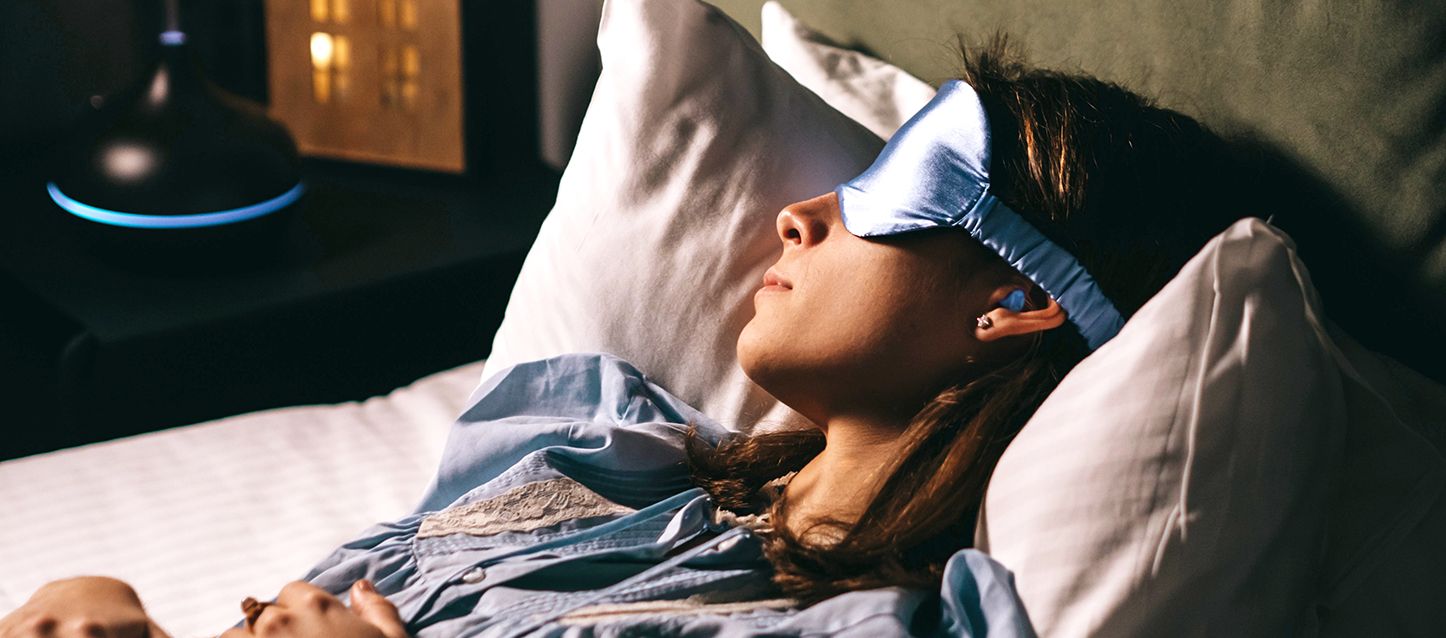 dormir con tapones chica de pelo castaño durmiendo con antifaz y pijama azul boca arriba y con tapones en los oidos