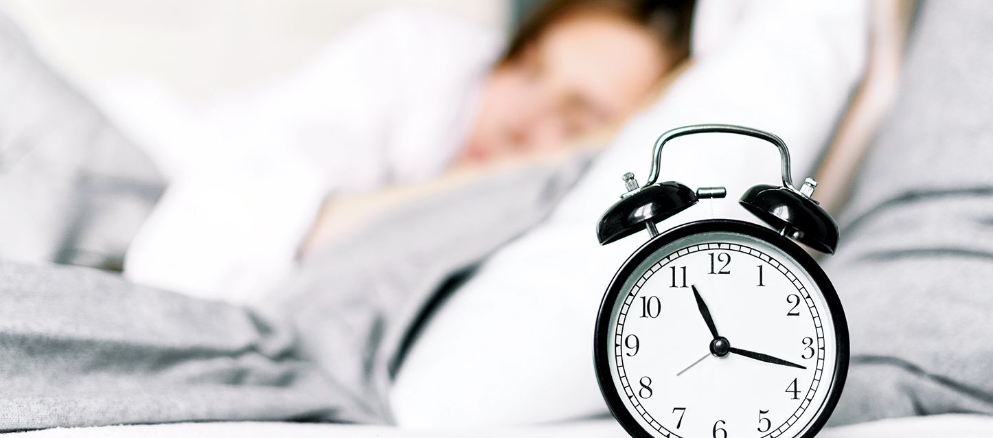 Cómo modificar nuestro horario de sueño reloj despertador antiguo de color blanco y negro en la mesita de noche y una chica durmiendo detrás