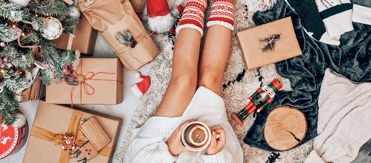 chica con jeresey de punto blanco sentada en el suelo con una taza de infusión en sus manos junto a un arbol de navidad con varios regalos y decoración navideña