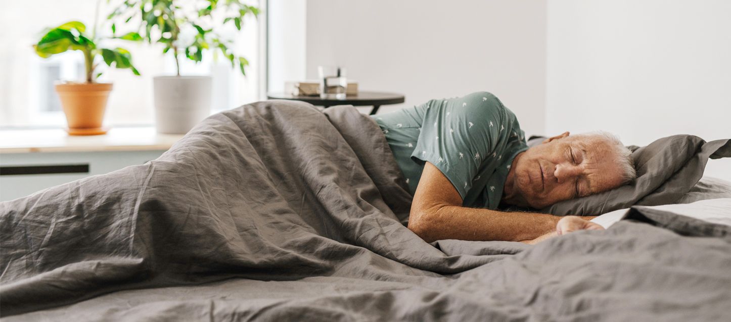 elegir un colchón para personas mayores hombre mayor con pijama gris durmiendo de lado tapado con edredon gris