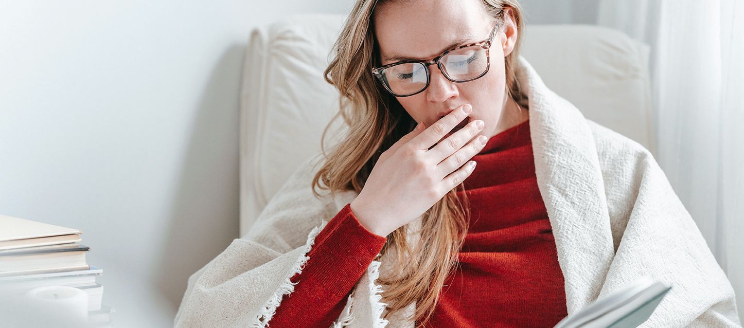 Cómo afecta a tu cuerpo trabajar de noche y dormir de día choca de pelo rubio largo con jersey rojo y tapada con una manta fina blanca por los hombros leyendo un libro y bostezando con la mano cubriendo su boca