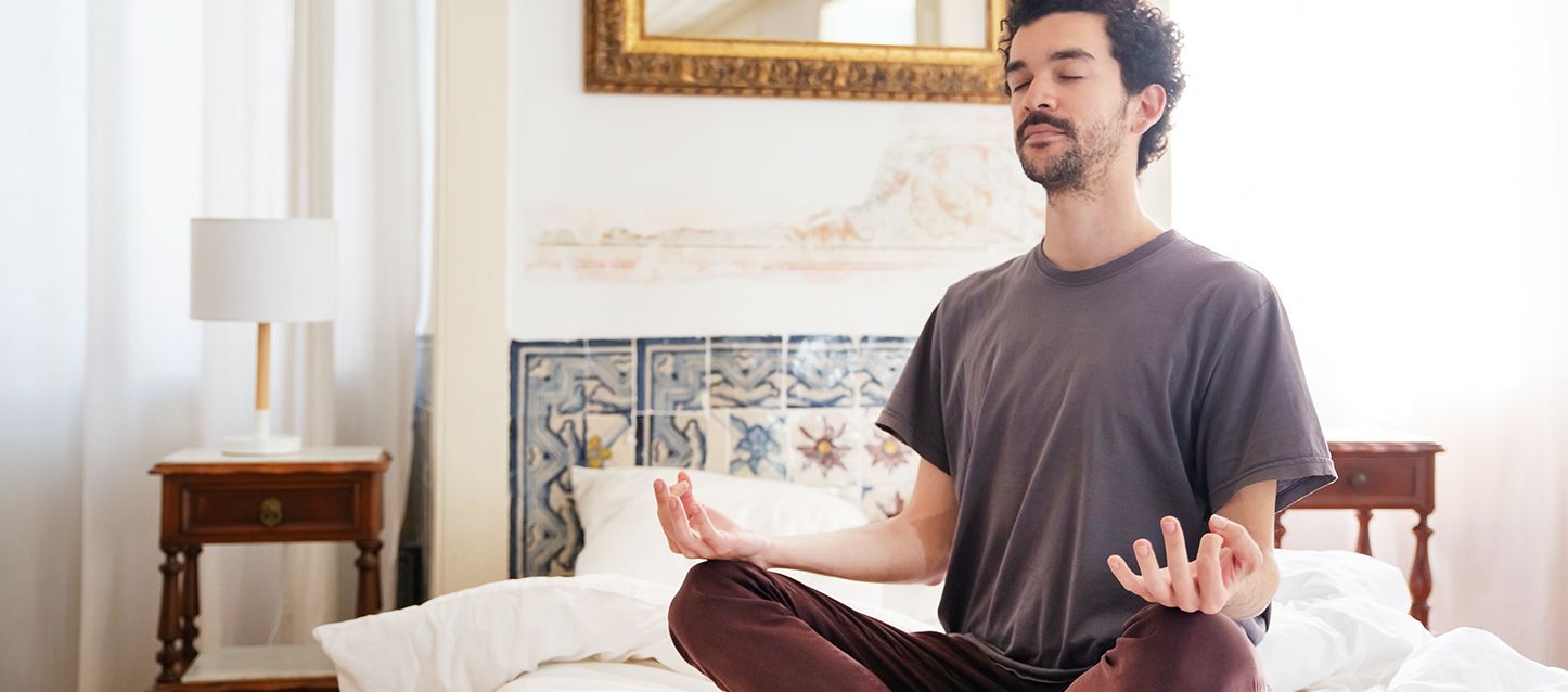 posturas que te relajan antes de irte a la cama chico moreno de pelo corto con camiseta gris y pantalones granates sentado en posición de meditación en el centro de su cama
