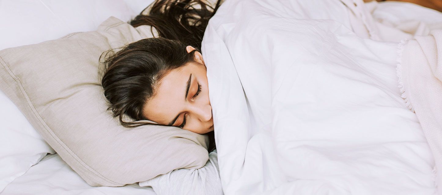 ¿por qué necesitamos dormir tanto? chica de pelo largo moreno durmiendo de lado en la cama tapada con el edredon de color blanco