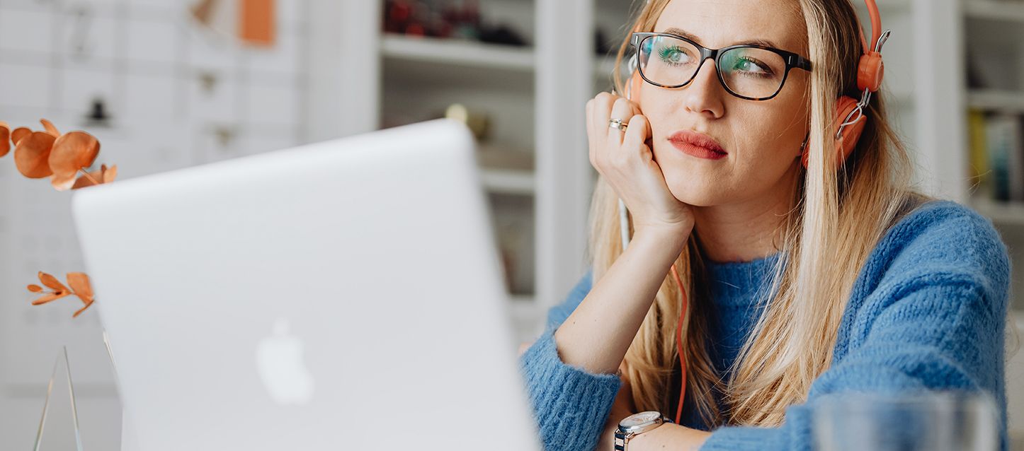 chica de pelo largo rubio, jersey azul, gafas y auriculares frente al laptop mirando pensativa