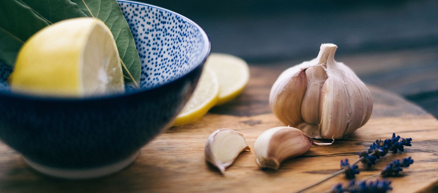 ¿Por qué poner un ajo debajo de la almohada? cabeza de ajo con dos dientes sobre la mesa, 2 ramas de lavanda, cuenco con limon y sus hojas y dos rodajas de limón sobre una tabla de madera