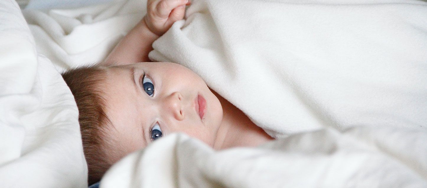 niño de pelo castaño con ojos azules tapado con una mantita blanca mirando