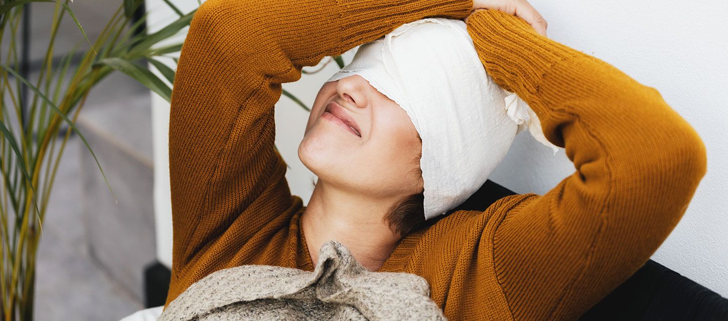 mujer con jersey marron claro de invierno con una venda que le cubre toda la cabeza y la cara hasta la nariz, con los brazos arriba de la cabeza y gesto de dolor.