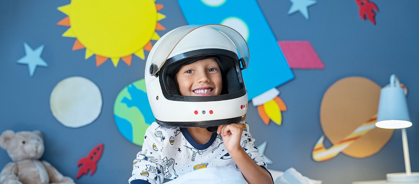 habitaciones infantiles divertidas niño con un casco de moto grande en una habitacion infantil divertida con la pared repleta de planetas y cohetes de decoracion