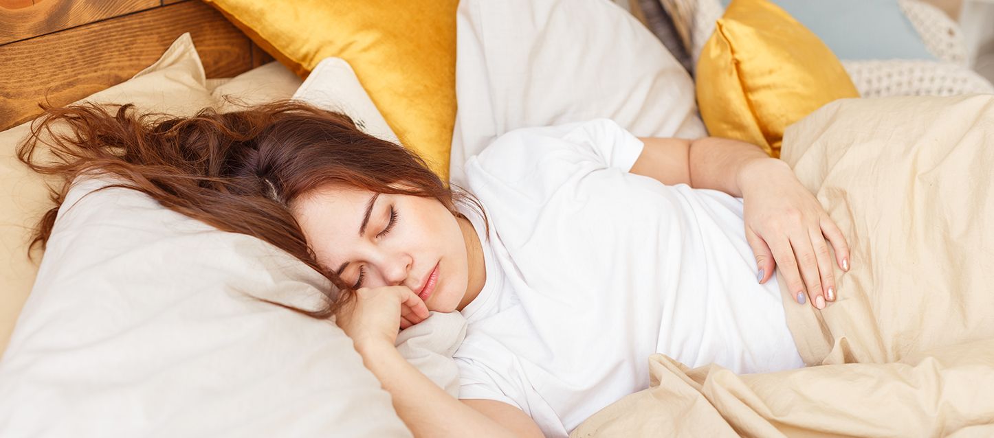 ¿Es bueno dormir con varias almohadas? chica de pelo largo castaño, camiseta blanca durmiendo con varias almohadas y tapada con camiseta beige