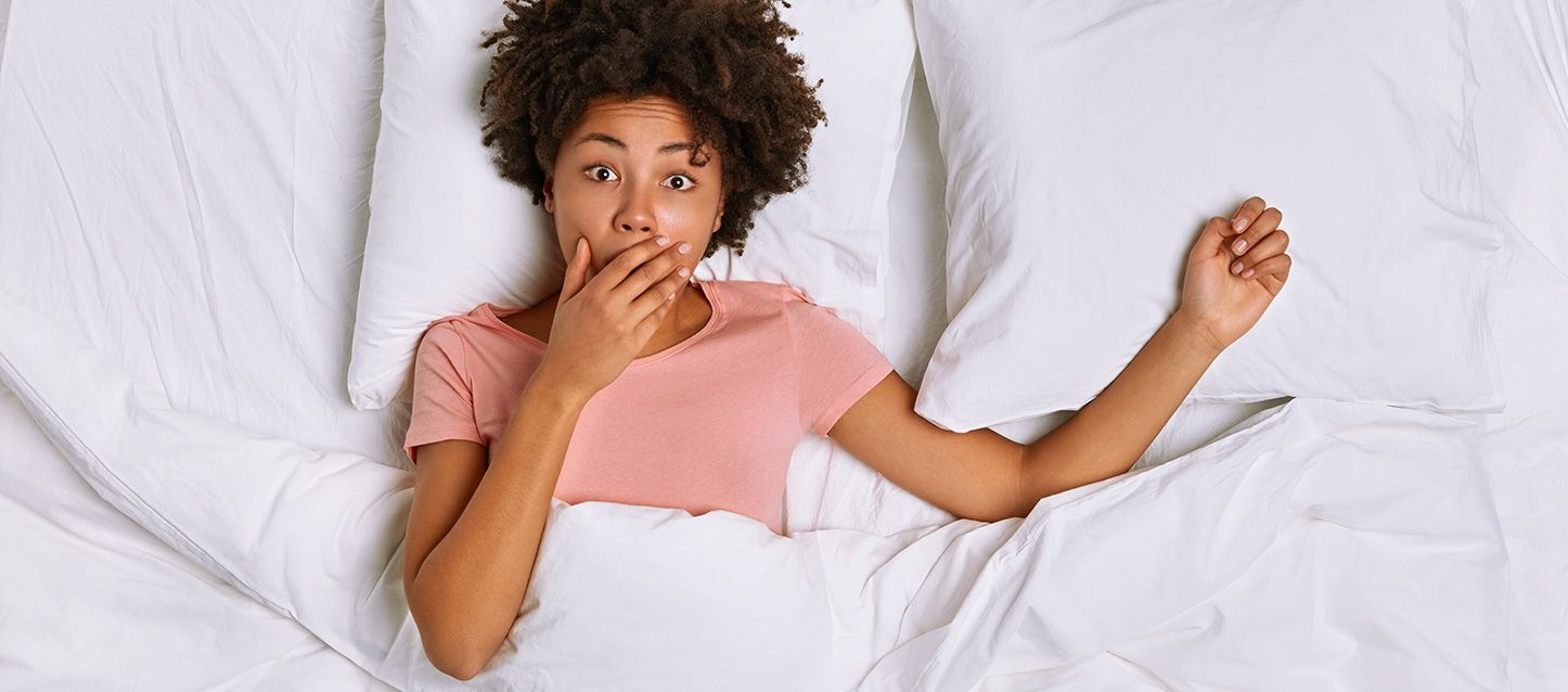 Qué es la somniloquía y cuáles son sus causas chica morena de pelo rizado, pijama rosa dentro de la cama tapada con edredon blanco con la mano en la boca y gesto de sorpresa.