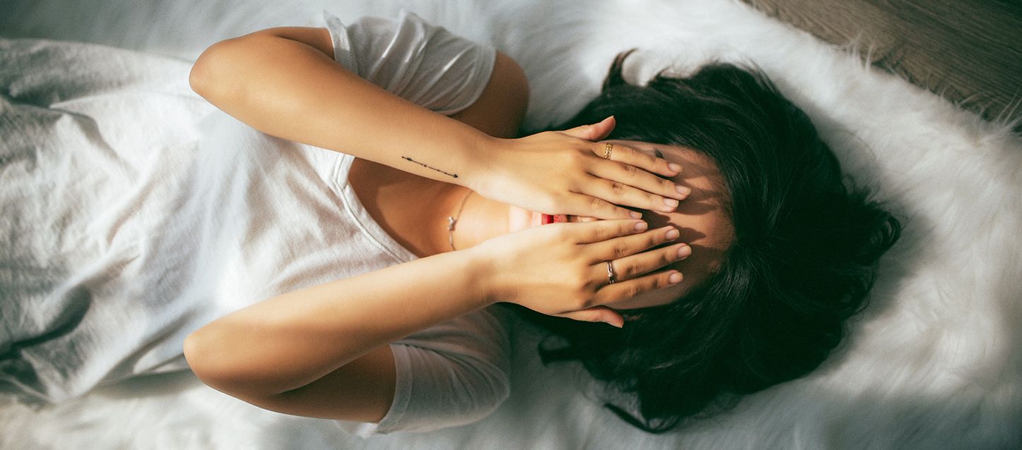Qué significa morir en un sueño chica de pelo negro tumbada boca arriba en la cama con camiseta blanca tapandose la cara con las manos asustada por soñar que se muere