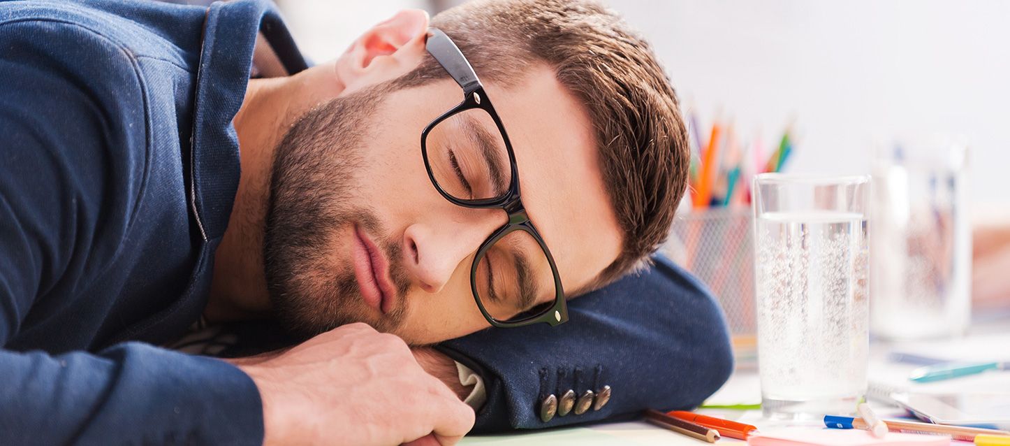 tratamientos naturales para la hipersomnia chico con barba y gafas durmiendo en una mesa junto a un vaso de agua y lapices en la mesa