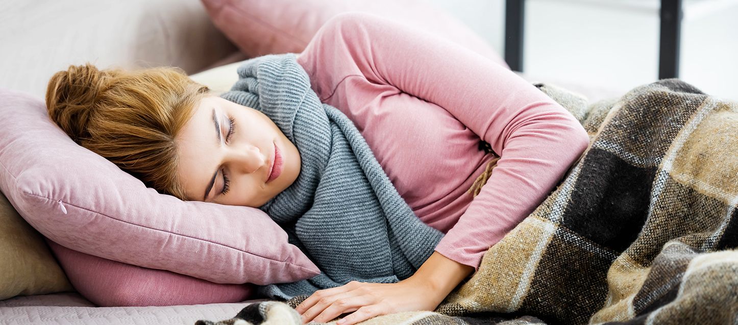 Escalofríos al dormir mujer de pelo castaño recogido con camiseta rosa, bufanda gris y tapada con manta a cuadros