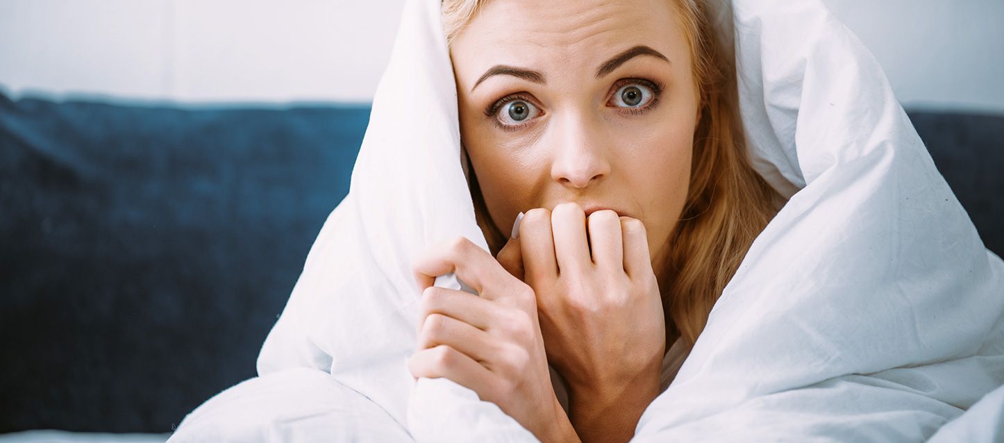 chica rubia de pelo largo y ojos verdes tapada con el edredon hasta la cabeza mordiendose la uñas con cara de susto