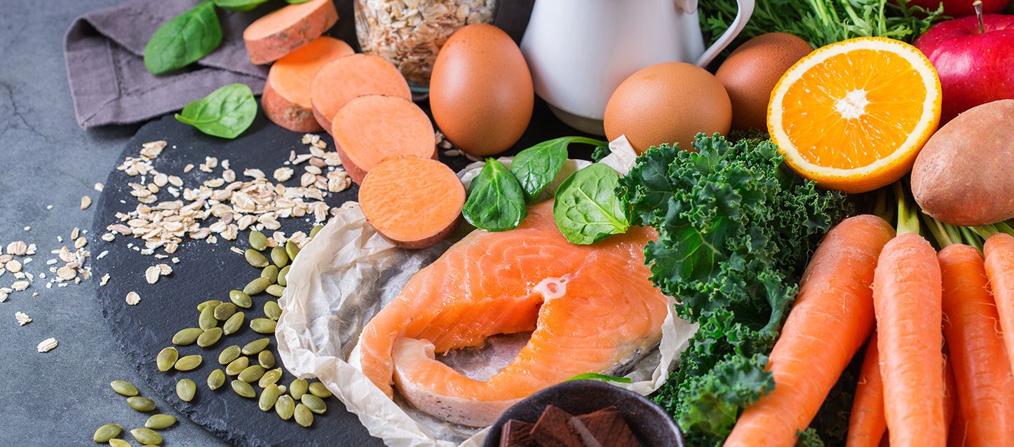Vitamina D mejora la calidad del sueño alimentos ricos en vitamina D sobre un plato llano de pizarra como boniatos, salmon, zanahorias, naranja, kale, huevos