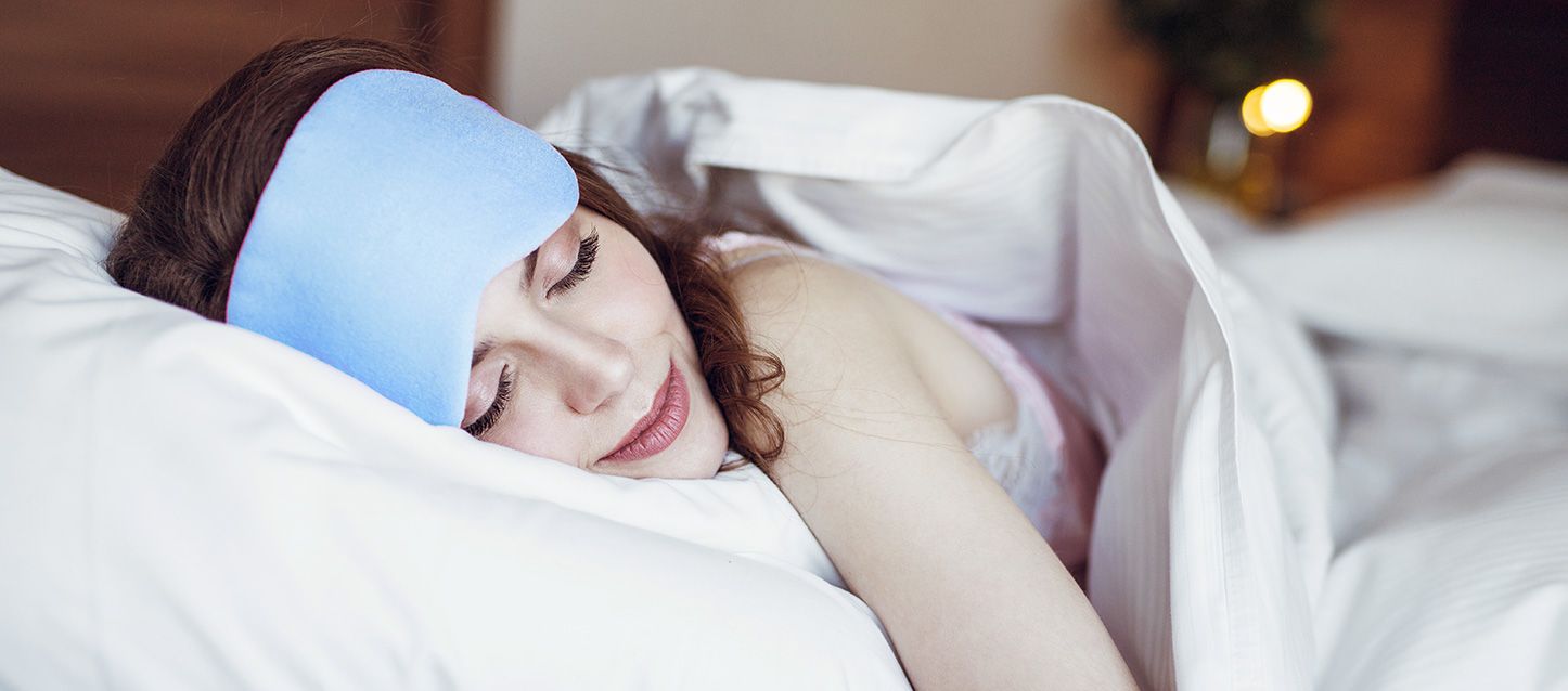 Sueño profundo mujer durmiendo boca abajo con antifaz en la frente azul y tapada con sábanas blancas