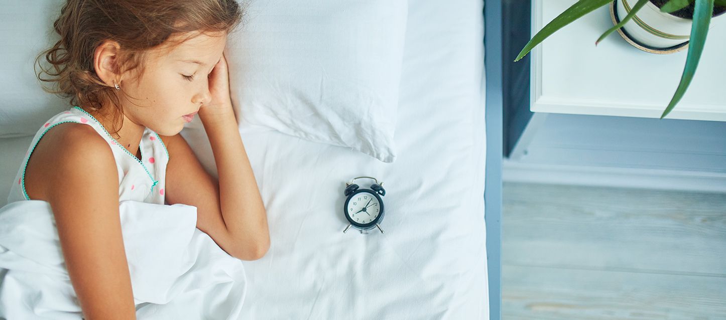 niña pequeña durmiendo de lado en la cama tapada con el edredón junto a un despertador