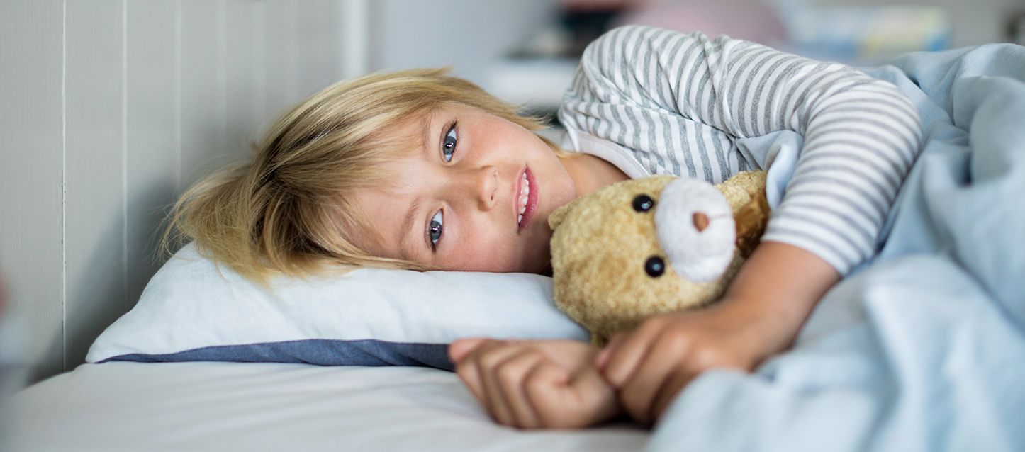 blog-1 niño pequeño rubia de ojos verdes acostado de lado en la cama abrazando a un osito de peluche mirando hacia la camara