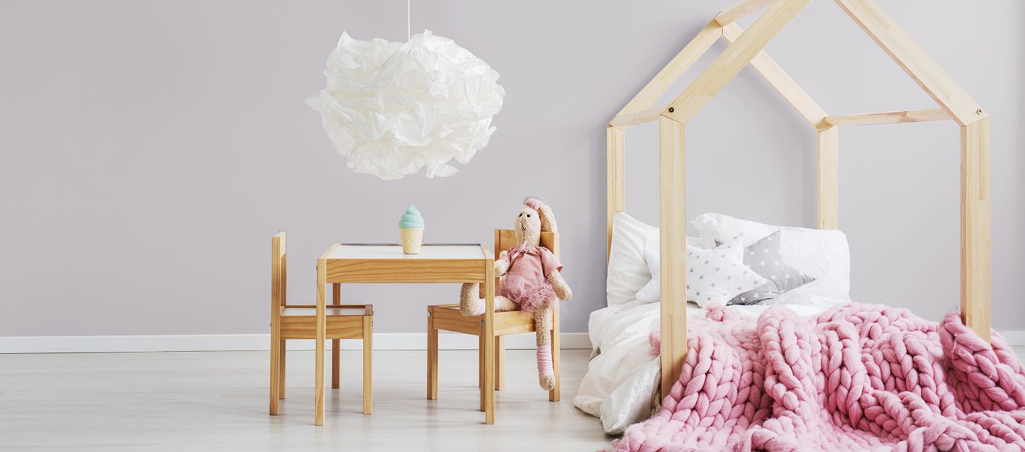 camas Montessori con ederedon rosa en una habitación con una mesa con sillas de madera y un peluche sentado en una silla con una lampara blanca