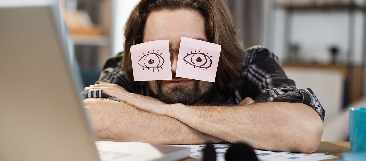 Chico dormido en frente del ordenador con 2 post it sobre el cristal de sus gafas con ojos pintados