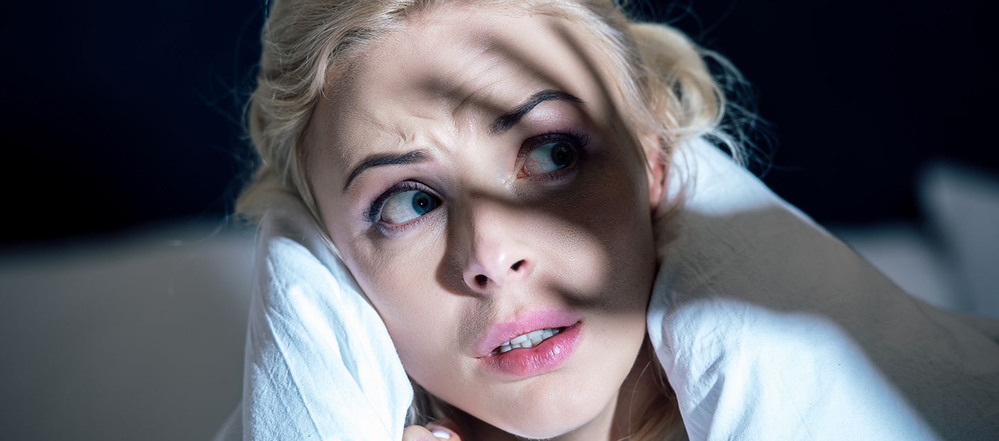Mujer rubia tapada hasta el cuello con el edredón blanco con cara de aterrorizada mirando hacia atrás