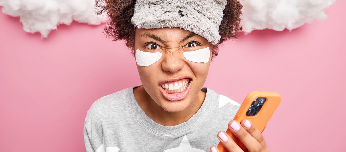 Chica morena con parches de belleza bajo los ojos sujetando un móvil en la mano rechinando los dientes