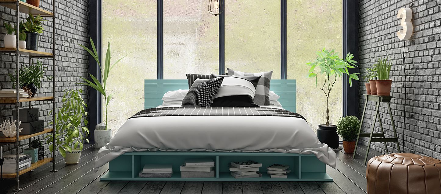 Habitación con suelo de madera y paredes de ladrillo. Colchón sobre soporte de madera con varios cojines y almohadas, pequeño macetero a la derecha y puf