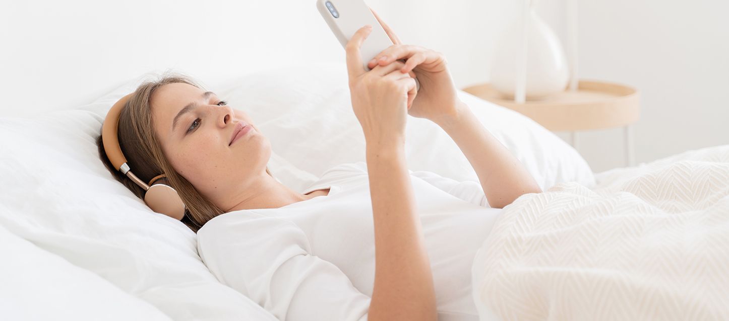 chica metida en la cama boca arriba con sabas, edredon y pijama blanco, con auriculares, mirando el movil escuchando asmr