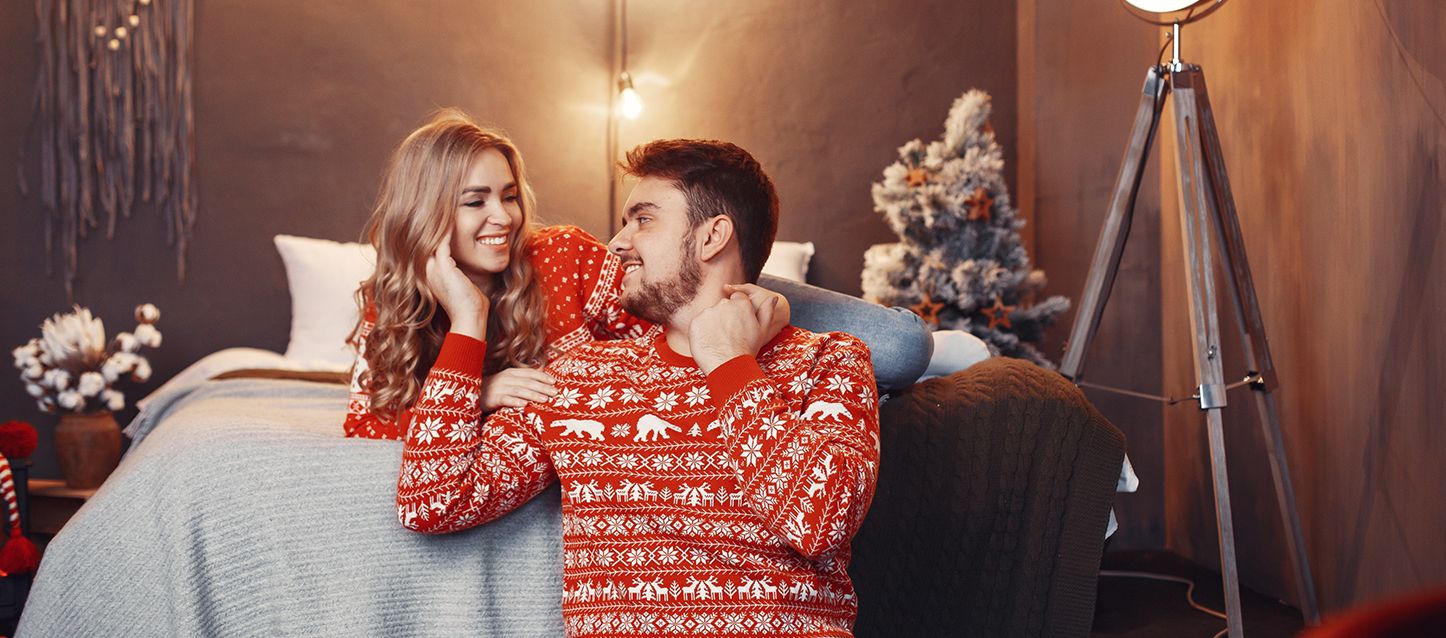 Cómo preparar el dormitorio para el invierno Pareja heterosexual con jerseys navideños apoyados en una cama mirándose felices
