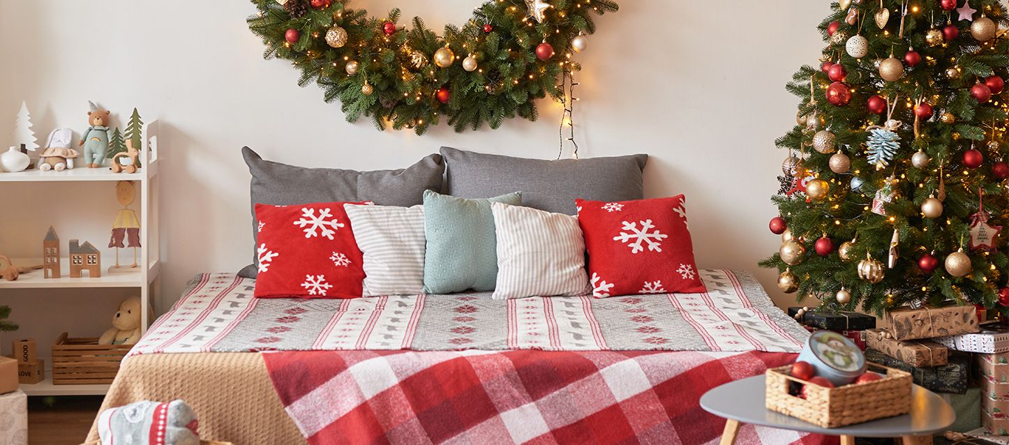 Dormitorio de invierno con una cama con mantas y cojines de motivos invernales, árbol de navidad a la derecha y estanteria con juguetes de madera a la izquierda