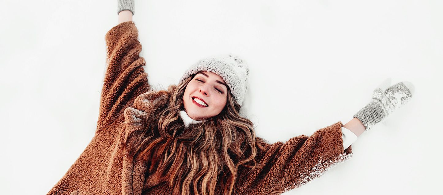 Mujer con una abrigo de invierno marrón, guantes y gorro gris tumbada boca arriba con los brazos extendidos sobre la nieve