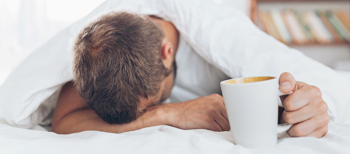 Cómo se puede mejorar el malestar matutino Hombre en la cama boca abajo cansado y con malestar sujetando una taza blanca en la mano