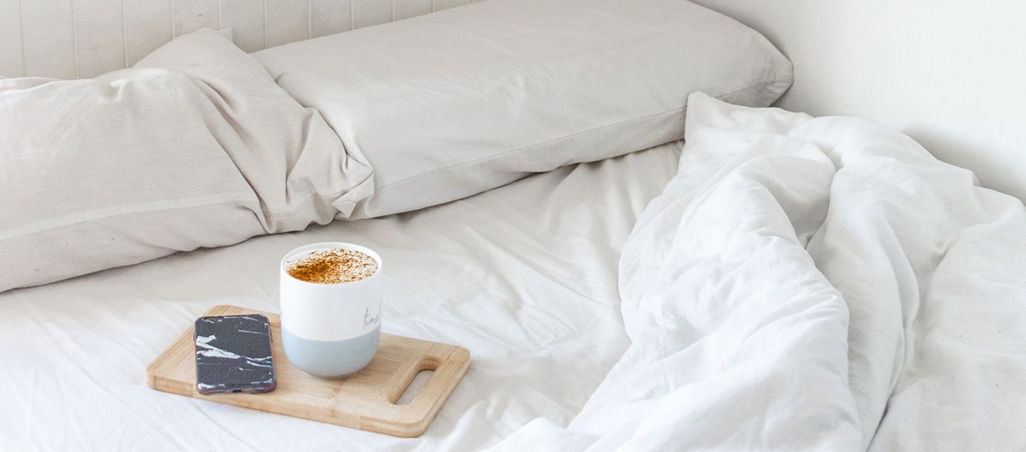 Cama con almohada y edredon con un capuccino y un móvil sobre una tabla de madera